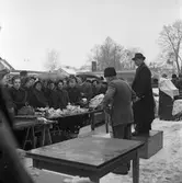 Auktion på Hamntorget, utomhus. Auktionsförrättaren står på en låda. Han har hatt och överrock på sig och klubban i handen. En man i krimmelmössa visar föremålen. På bordet framför dem står porslin och andra småsaker. Längst till höger står möbler, täckta mot väder och vind. Publiken består mest av kvinnor. Några har paraply. I bakgrunden syns Texacomacken.