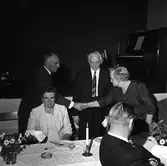 Axel Karlsson, Medåker, får gåva, är Reinhold Carlssons text till denna bild.
En man och en kvinna tar varandra i hand. Mannen mellan dem är Axel. En kvinna och en man sitter vid ett bord. De har just slutat äta. Bordet är dukat med blombuketter, ljusstake och askfat. På scenen, i bakgrunden, står ett piano. Personerna är uppklädda.