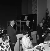 Axel Karlsson, Medåker, har fått motta en gåva. En äldre man håller en liten ask i handen.
Uppklädda människor, bordet är dukat och det står blomvaser på bordet och på scenen. På scenen finns dessutom ett piano.
Bilden är möjligen tagen i Medåkers bygdegård.