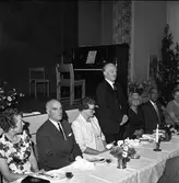 Axel Karlsson, Medåker, håller tacktal.
Uppklädda människor sitter vid långbord. Det står blombuketter på bordet.
Bilden är möjligen tagen i Medåkers bygdegård.
På scenen, bakom dem, står ett piano.
