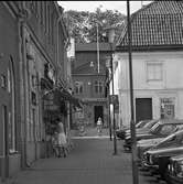 Västra sidan av Stora torget. En kvinna parkerar sin cykel mellan Frisören och café Saga. Två flickor med var sin barnvagn kommer gående i Baggens gränd. Bakom dem ses Stadskällaren på Nygatan. Till höger i bild ligger Duka-butiken och flera bilar står parkerade.