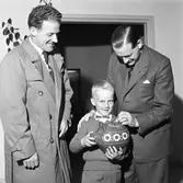 Bankdirektör S E Zakrisson, från Sparbanken, belönar en liten sparare. Han stoppar en slant i spargrisen.
Bankdirektören i kostym, en liten pojke och möjligen pojkens pappa, iklädd överrock.