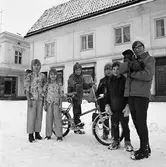 Barn med julklappar. Två flickor i nya kläder. En pojke med en minicykel. Flickan längst till höger har en hund i famnen.
Butiken i bakgrunden har REA.