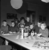 Barndaghemmet. Sju barn och fröken Astrid Eriksson, sitter vid ett bord. Där finns pussel och barnen leker med bilar och båtar.