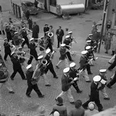 Barnens Dag firas. En blåsorkester marscherar i paraden. Människor står på trottoaren och ser på. I bakgrunden ses Shells bensinstation. Platsen är Jädersvägen och Jäderstullen.