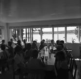 Mellanmål vid Barnens Dags koloni i Högsjön, väster om Arboga. De många fönstren erbjuder en vacker utsikt, över sjön. Barnen sitter vid två långbord och dricker mjölk och äter knäckebröd.