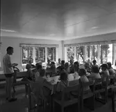 Barnens Dagskolonin vid Högsjön, väster om Arboga. Barnen sitter vid två långbord, dricker mjölk och äter knäckebröd. En ledare, iklädd shorts, berättar.