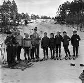 Barnskidskolan har avslutning. Elva barn och en lärare är uppställda nedanför backen. De har längdskidor på sig medan stavarna är satta i snön. De är klädda i stickade tröjor och mössor eller i anorack. De har fått var sitt äpple.