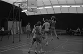 Herrlag spelar basket i plasthallen. Några personer sitter på bänkar och tittar på.