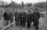 Herrar i vit skjorta och slips ser på byggplatsen för det nya badet i Ekbacken. 
Utomhusbassängen håller på att grävas ur.
Två av männen är identifierade: Den sjätte mannen från vänster är Hildning Lund och den åttonde är Nils Brodin.
Till vänster i bild ses några ungdomar som sysslar med byggnationen.