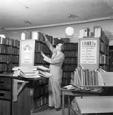 Bibliotekarie Östen Bengtsson.
Biblioteket ligger på Nygatan (i gamla Gästis).
Kostymklädd man som sätter upp böcker i bokhylla, omgiven av bokhyllor.