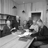 Biblioteket på det nya ålderdomshemmet Strömsborg. Män och kvinnor samlade runt ett bord med böcker. Kvinnan i prickig klänning är Ellen Sjöstam.
Bokhyllor ses efter väggen.