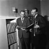 Arboga Blåsorkester repeterar
En man plockar fram noter, mannen bredvid håller i en trumpet
Vid sidan av dem står en hylla men noter.