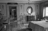 Bostadsinteriör, Crugska gården. Sovrum med två sängar. En chiffonjé med en stol framför, en byrå med en spegel över. Väggarna är dekorerade. Ett fönster. En ljuskrona i taket.
Genom dörröppningen kan man se in i flera rum.