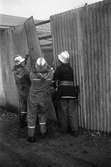 Det brinner i en verkstad på Norra Ågatan. Tre brandmän lossar en plåt för att kunna ta sig in. De är klädda i hjälm och skyddskläder.