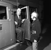 Brandkåren avtackar veteranen, brandmästare Erik Jansson. 
Kostymklädd man som sitter i brandbil. Dörren öppnas av en uniformsklädd brandman med hjälm.