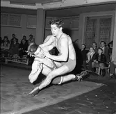 Brottningsmatch mellan AAK och Sala.
Publiken sitter på bänkar och ser på de tävlande.
AAK = Arboga AtletKlubb