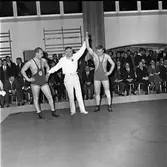 Tävlingen Fyrstadsbrottning mellan Arboga, Eslöv, Uddevalla och Falkenberg.
Arboga Atletklubbs Roland Andersson går segrande ur sin match.
Publiken sitter efter väggarna i gymnastiksalen.