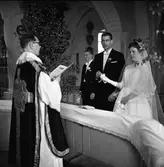 Ernst Hansson förrättar vigsel i Säterbo kyrka. Herr Olsson och fröken Tallqvist avlägger sina löften. Bruden har tagit av handsken på den hand där vigselringen ska vara.
Kyrkan är dekorerad med björkar.