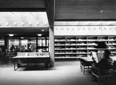 Arboga Stadsbibliotek, interiör. Entrén till biblioteket ses till vänster. Till höger står hyllorna med tidskrifter. En man sitter och läser en dagstidning vid det stora bordet. De stora fönstren, mot Arbogaån, ligger utanför bilden, till höger.