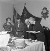 Elektrikerförbundets Arbogaavdelning har jubileumsfest. Gående bord. Två kvinnor och två män tar för sig av maten. På väggen hänger två fanor och en pendyl.