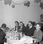 Elektrikerförbundets Arbogaavdelning har jubileumsfest. Män och kvinnor som just avslutat en måltid. Bordet är dukat med en ljusstake och en vas med en blomma. Väggdekorationer.
Kvinnan, vid bordet bakom, är fru Bergström.
