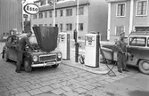 Bensinstationen ESSO på Nygatan. Mannen till vänster heter Andersson och bor i Götlunda. Han är duktig på skidor och motocross. Mannen som tankar bilen kallas Celle.
Här finns två bensinpumpar. Männen är klädda i overaller.
Intill macken ligger ett konditori.