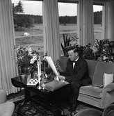 Erik Holm (kallad Glass-Holm), sitter i sitt vardagsrum och tittar på sina 50-årspresenter. Han har bland annat fått ljusstakar och många blommor.
Möblemanget består av soffa, två fåtöljer, ett bord och en ryamatta. Stora fönster vetter mot landskapet utanför.