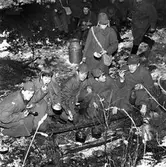 FBU-ungdomar övar i skogen. Det är vinter och snö. Alla är klädda i yllerockar och mössor med öronlappar eller båtmössor. En ledare, i skärmmössa, sitter intill eldstaden där något kokar i grytorna. 
Frivillig Befälsutbildning