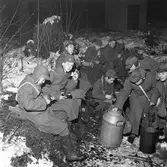 FBU-ungdomar övar. 
Unga pojkar sitter på granris och äter. De är klädda i yllerockar och mössor med öronlappar. Det är vinter och snö. De äter ur sina plåtburkar. En stor mjölkkanna är placerad framför dem.
FBU betyder Frivillig befälsutbildning