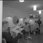 Frisör Axel Fritiof Strandberg i frisersalongen på det nya ålderdomshemmet Strömsborg. Hans ordinarie hårsalong ligger på Stora torget.
Atschy klipper en man. En kvinna sitter i en torkhuv. En man läser en tidning. Det är speglar på väggen.