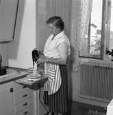 Fru Blomberg i köket. Hon är iklädd ett förkläde och har just tagit fram en skål och en el-visp. Fönstret är öppet och hon har myggnätet isatt. En krukväxt står på fönsterbrädan.
Kvinnan är gift med Jakob Blomberg som arbetar på Arboga Mekaniska Verkstad.
AMV