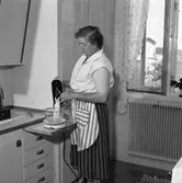 Fru Blomberg i köket. Hon är iklädd ett förkläde och har just tagit fram en skål och en el-visp. Fönstret är öppet och hon har myggnätet isatt. En krukväxt står på fönsterbrädan.
Kvinnan är gift med Jakob Blomberg som arbetar på Arboga Mekaniska Verkstad.
AMV