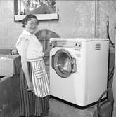 Fru Blomberg (hustru till Jakob Blomberg på Arboga Mekaniska Verkstad) visar en tvättmaskin.
Kvinna, iklädd förkläde, står i källaren intill en ny tvättmaskin.
AMV