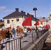 Första maj. Demonstrationståg på Kapellbron. Per-Olov Nilsson, i brun jacka, bär ett plakat.
Kapellbron är dekorerad med röda fanor. Människor bär plakat.
I bakgrunden ses kvarteret Stadsgården.
