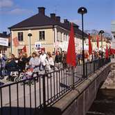 Första maj. Demonstrationståg på Kapellbron. Människor bär plakat. Bron är dekorerad med röda fanor.
I bakgrunden ses kvarteret Stadsgården.
Socialdemokrater.