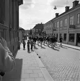 Första maj. Demonstration. Arboga Blåsorkester går i täten. De är i jämhöjd med kvarteret Stadsgården. Mariannes Klippotek, Jeansshoppen och en damfrisering ligger i byggnaden. Demonstrationståget är på väg över Kapellbron, upp mot centrum. Fanor och plakat syns bakom musikkåren.
Socialdemokrater