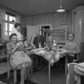 Försäljning hos pensionärerna på Strömsborgs äldreboende. En man, fyra kvinnor och en kvinnlig personal. På bordet står diverse hantverk. I bakgrunden ses en vävstol och en kakelugn.