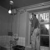 Gamla ålderdomshemmet rustas. En man, iklädd keps och arbetsbyxor, står i fönstret. Renovering av hygienutrymme. Disko, kranar, rörledningar. En naken glödlampa i taket.
