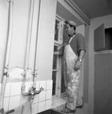 Gamla ålderdomshemmet rustas. En man, iklädd keps och arbetsbyxor, står i fönstret. Renovering av hygienutrymme. Diskho, kranar och rörledningar. En naken glödlampa i taket.
