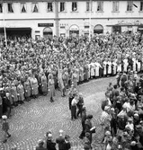 En stor publik har samlats på Stora torget. Konung Gustaf Vl Adolf gästar Arboga under sin Eriksgata. I främsta ledet står Arboga Lottakår och vid sidan av dem, kvinnor i folkdräkt.
I bakgrunden ses Lundborgska fastigheten med Sture-Bio, Tidstrands garn & trikå, skoaffären, Strandbergs och Konditori Saga.
För värdskapet svarade stadsfullmäktiges ordförande Jonas Carlsson och kommunalborgmästare Danliel Ekelund. Den organisationskommitté som ansvarade för arrangemangen, hade hos drätselkammaren begärt en summa av 3000 kronor för att täcka kostnaderna vid kungabesöket. Beloppet beviljades. 
Tiden för kungens besök var beräknad till 130 minuter.