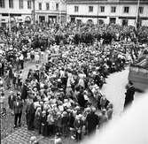 Åhörare har samlats på Stora torget för att lyssna på Kung Gustaf Vl Adolf. Konungen besöker Arboga i samband med sin Eriksgata. Barn, med flaggor i händerna, har samlats nära scenen. Åt höger, i bild, ses Frälsningsarmén. 
Järnbolaget, Speceriaffären, Haglunds Guld och Eklöws Bosättning ses i bakgrunden.
För värdskapet svarade stadsfullmäktiges ordförande Jonas Carlsson och kommunalborgmästare Danliel Ekelund. Den organisationskommitté som ansvarade för arrangemangen, hade hos drätselkammaren begärt en summa av 3000 kronor för att täcka kostnaderna vid kungabesöket. Beloppet beviljades. 
Tiden för kungens besök var beräknad till 130 minuter.