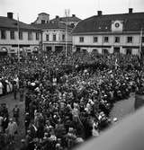 Publik på Stora torget. Skolbarn med flaggor, Frälsningsarmén, unga och gamla har samlats för att se och höra kung Gustaf Vl Adolf. Han gästar Arboga under sin Eriksgata. Han står på den uppbyggda scenen till höger, strax utanför bild.
Järnbolaget, Specerier, Haglunds Guld och Eklöws bosättning ses i bakgrunden.
För värdskapet svarade stadsfullmäktiges ordförande Jonas Carlsson och kommunalborgmästare Danliel Ekelund. Den organisationskommitté som ansvarade för arrangemangen, hade hos drätselkammaren begärt en summa av 3000 kronor för att täcka kostnaderna vid kungabesöket. Beloppet beviljades. 
Tiden för kungens besök var beräknad till 130 minuter.