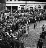 En stor publik har samlats på torget. Kung Gustaf Vl Adolf ska tala från en uppbyggd scen intill rådhuset. Han gästar Arboga under sin Eriksgata. I åhörarleden ses Arboga Hemvärnskår och Arboga Lottakår.
I bakgrunden ses Sture-bio, Tidstrands garn, Skoaffären och Atschy Strandbergs frisörsalong. 
För värdskapet svarade stadsfullmäktiges ordförande Jonas Carlsson och kommunalborgmästare Danliel Ekelund. Den organisationskommitté som ansvarade för arrangemangen, hade hos drätselkammaren begärt en summa av 3000 kronor för att täcka kostnaderna vid kungabesöket. Beloppet beviljades. 
Tiden för kungens besök var beräknad till 130 minuter.