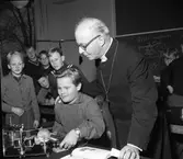 Nybyggda Gäddgårdsskolan har besök av biskop John Cullberg. Börje Hagström gör ett experiment på fysiklektionen och biskopen ser intresserat på. I bakgrunden ses klasskamraterna Örjan Carlén (i knäppetröja), okänd som kikar fram under kompisarna, längst bak från vänster, okänd, Karl-Ivar Pettersson och Bernt Mellblom. Bakom ryggen på biskopen skymtar Reijo Koivisto.
