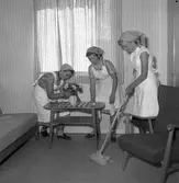 Hemkunskapsundervisning på Gäddgårdsskolan. Anita Bengtsson ordnar med blommorna, Sonja Berglind torkar bordet och en okänd flicka skurar golvet. Alla tre är klädda i sjaletter och förkläden.