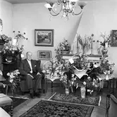 Hemma hos byggmästare Johan Sörén på bemärkelsedagen
En man sitter i en fåtölj i sitt vardagsrum. På chiffonjén står blommor i vaser, på spiselkransen och på golvet står korgar och vaser med blomsteruppsättningar och buketter.