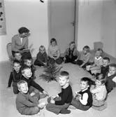 HSB´s barnträdgård. Sagostund. Fröken läser ur en bok och barnen sitter i ring på golvet. Mitt i ringen står en liten julgran.
(HSB betyder Hyresgästernas sparkasse- och byggnadsförening)