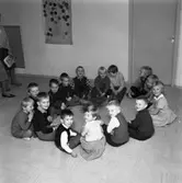 HSB´s barnträdgård. Barnen sitter i ring på golvet. Mitt i ringen står en liten julgran. Fröken anas till vänster i bild.
(HSB betyder Hyresgästernas sparkasse- och byggnadsförening)