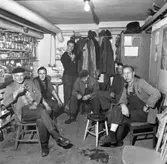 Rökpaus i HSB´s verkstadslokaler. Från vänster: Erik Andersson, Folke Ivansson, Bertil Jonsson, Andersson, Nils Persson och Erik Persson.
Sex män har rast i källarlokalen. Fyra röker pipa, en cigarett och är passiv rökare. De har arbetskläder på sig, skyddsrockar och blåkläder. Efter ena väggen har de glasburkar med skruvar och andra småprylar i. En hatthylla är full av jackor och mössor. I hörnet finns ett handfat. En naken glödlampa lyser i taket.
(HSB betyder Hyresgästernas sparkasse- och byggnadsförening).